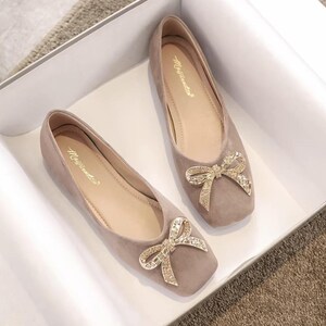 May include: A pair of taupe-colored flats with square toes, each adorned with a sparkling bow embellishment. The shoes are displayed inside a white box, showcasing their elegant design and style.