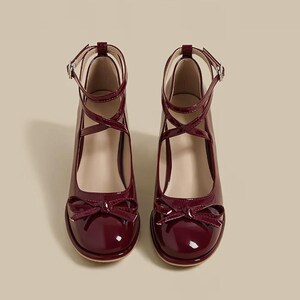 May include: A pair of burgundy patent leather Mary Jane style shoes. The shoes have a rounded toe, a bow detail, and a criss-cross ankle strap with a buckle. The heels are approximately 2 inches high.