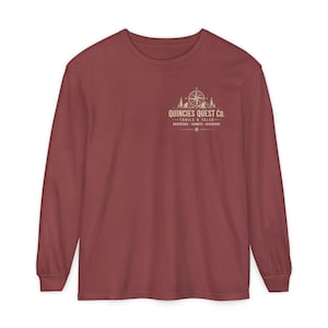 Op de afbeelding: Een shirt met lange mouwen en een ronde hals in een gedempte baksteenrode kleur. Op de linkerborst staat een grafische print met een kompas, bergsilhouetten en de tekst "QUINCIES QUEST CO. TRAILS & TALES ADVENTURE GROWTH DISCOVERY".