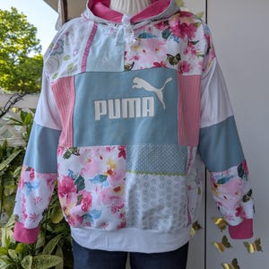 Könnte beinhalten: Ein Patchwork-Kapuzenpullover aus einer Mischung aus floralen, gepunkteten und einfarbigen Stoffbahnen in Rosa, Weiß und Hellblau. Auf der Brust befindet sich ein weißes Puma-Logo mit Schriftzug. Das Kleidungsstück hat eine rosa gefütterte Kapuze mit weißen Kordeln.