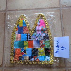 Könnte beinhalten: Ein gesteppter Stoffaufnäher mit spitzer Oberkante, bestehend aus einem bunten Patchwork aus verschiedenen Mustern und Farben, eingefasst mit goldener Paillettenborte. Der Artikel ist in einer Klarsichtfolie mit einem Zettel Modell 4 verpackt.