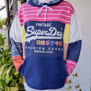 Könnte beinhalten: Ein Vintage Superdry Hoodie mit Colorblock-Design. Der Hoodie hat rosa und weiße horizontale Streifen, eine marineblaue Tasche und Kapuze sowie das Superdry-Logo. Die Ärmel haben ein Patchwork-Design mit blau-weißen Mustern.