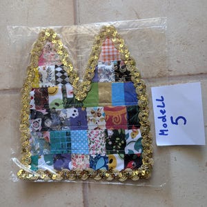 Könnte beinhalten: Ein stoffaufnäher in Form von Katzenohren mit einem Patchwork-Design aus verschiedenen bunten Mustern, darunter Blumen, Karos und grafische Drucke. Die Ränder sind mit goldenen Pailletten verziert. Ein kleiner weißer Zettel mit dem Text Modell 5 liegt bei.