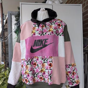 Könnte beinhalten: Ein Patchwork-Hoodie mit floralem Muster, rosa, weißen und olivgrünen Einsätzen. Auf der Vorderseite befindet sich ein großes rosa Panel mit dem schwarzen Nike-Logo. Die Kapuze ist schwarz mit Kordelzug.