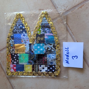 Könnte beinhalten: Ein stoffaufnäher in Katzenohrform mit einem Patchwork-Design aus verschiedenen blau, schwarz, weiß und gold gemusterten Quadraten. Die Ränder sind mit goldenen Pailletten besetzt. Ein weißer Zettel daneben trägt den handschriftlichen Text Modell 3.
