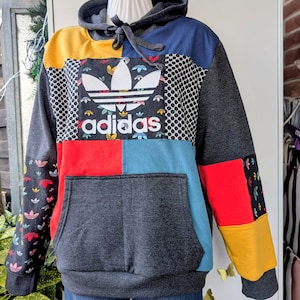 Könnte beinhalten: Ein farblich abgesetzter Kapuzenpullover mit dunkelgrauer Basis und Einsätzen in Senfgelb, Blau und Rot. Die Vorderseite zeigt ein großes weißes Adidas-Trefoil-Logo und Schriftzug auf einem schwarz gemusterten Bereich. Mit Kängurutasche.