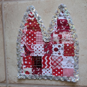 Könnte beinhalten: Ein katzenohrförmiges Stoffornament mit einem Patchwork-Design aus roten und weißen Quadraten mit verschiedenen Mustern wie Sternen, Ankern, Punkten und Streifen. Die Kanten sind mit einer schimmernden silbernen Paillettenborte verziert.