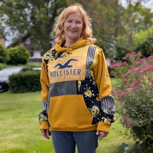 Könnte beinhalten: Ein senfgelber Kapuzenpullover mit Patchwork-Design, bestehend aus schwarz-weißen vertikalen Streifen und einem schwarzen Blumenmuster. Das Mittelteil zeigt das dunkelblaue Hollister California Logo und Schriftzug mit einer Kängurutasche.