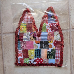 Könnte beinhalten: Ein katzenohrförmiger Stoffaufnäher mit einem Patchwork-Design aus verschiedenen bunten Mustern, darunter Blumen-, Vichy-, Punkte- und Smiley-Drucke. Die Ränder sind mit roten Pailletten verziert und der gesamte Artikel befindet sich in einer Klarsichtfolie.