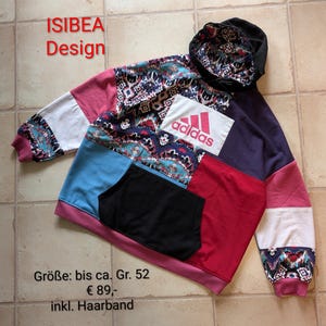 Könnte beinhalten: Ein mehrfarbiger Patchwork-Hoodie mit schwarzer Kapuze und Tasche. Das Design beinhaltet verschiedene Muster und Farben, mit einem weißen Adidas-Logo. Der Text "ISIBEA Design" ist rot.