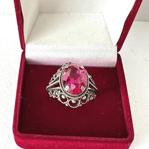 Può includere: Un anello in argento con una pietra preziosa rosa brillante dal taglio ovale, incastonata in una fascia decorativa in stile filigrana. L'anello è esposto all'interno di un portagioie quadrato foderato in velluto, con esterno rosso scuro.
