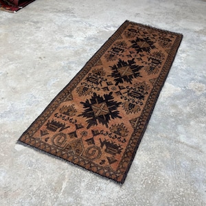 May include: A rectangular runner rug featuring a warm brown background with a repeating pattern of dark brown and black geometric motifs and stylized floral shapes. The rug has a dark border and short fringe along the narrow ends of the piece.