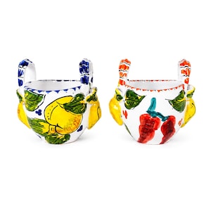 Può includere: Due tazze in ceramica con due manici ciascuna. La tazza a sinistra presenta limoni gialli e foglie verdi con manici a righe blu. La tazza a destra mostra peperoncini rossi e foglie verdi con manici a righe arancioni su uno sfondo bianco.