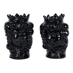 May include: A pair of glossy black ceramic vases shaped like human heads wearing crowns. Each vase features detailed facial features, textured hair, and a scalloped crown rim. The dark, reflective finish covers the entire surface of both vessels.