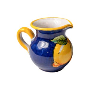 Puede incluir: Una jarra de cerámica con cuerpo azul, asa amarilla y borde amarillo. La jarra presenta un diseño de limón pintado a mano con hojas verdes. Es un artículo de cocina decorativo.
