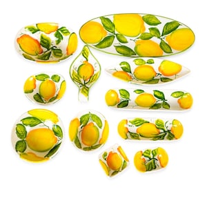 Puede incluir: Un conjunto de vajilla de cerámica blanca con limones amarillos y hojas verdes pintados a mano. El set incluye varios cuencos, fuentes ovaladas, un plato con forma de hoja y bandejas rectangulares, todos decorados con un motivo de limones.