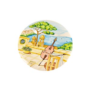Może przedstawiać: Okrągły, ręcznie malowany ceramiczny talerz przedstawiający scenę nadmorską z wiolonczelą, nutami i małym żółtym krzesłem. Talerz ma jasnoniebieskie niebo, zielone drzewa i widok na morze.