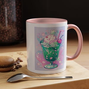 May include: White ceramic mug with a pink interior and handle. The mug features a colorful illustration of a green dessert with ice cream, a cherry, and a straw. The background has hearts and stars.