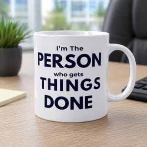 May include: A white ceramic mug with a handle, featuring dark blue text that reads: I'm The PERSON who gets THINGS DONE. The mug has a cylindrical shape and a smooth finish, designed for holding hot or cold beverages in a standard office setting.