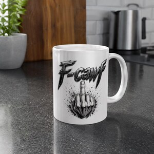 May include: A white ceramic mug featuring a black graphic illustration of a hand making a middle finger gesture. Above the hand, the text F-cawf is printed in a stylized, jagged black font. The design includes black ink splatter effects.