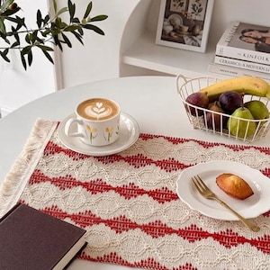 Puede incluir: Una mesa con un camino de mesa estampado en rojo y blanco, una taza de café con arte latte, un plato con un pastel y una cesta de frutas. Una foto enmarcada y libros están en un estante en el fondo.