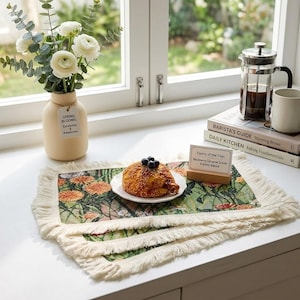May include: A still-life scene featuring a floral placemat with a pastry on a plate. A vase of white flowers and a coffee press are also present. The placemat has a fringed edge and a botanical design. A sign reads "Pastry of the Day: Blueberry Sesame Scone Freshly Baked."