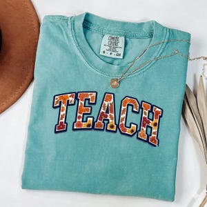 May include: A teal Comfort Colors t-shirt with the word "TEACH" in a collegiate-style font. The letters are filled with a floral pattern of orange, yellow, and red autumn leaves. A gold necklace with a pendant rests on the shirt.