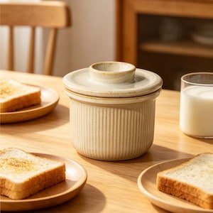 May include: A beige ceramic butter dish with a lid, featuring vertical ribbed detailing. The lid has a small, circular handle. Slices of toasted bread sit on wooden plates, alongside a glass of milk, creating a breakfast scene.