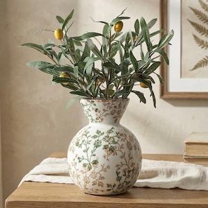 May include: A white ceramic vase with a floral pattern, holding olive branches with green leaves and yellow olives. The vase has a bulbous base and a flared top, and is placed on a wooden table.