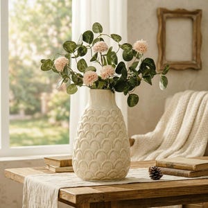 May include: A cream-colored, textured vase with a scalloped pattern holds pink flowers and green foliage. The vase sits on a wooden table with books and a pinecone. A window and a framed picture are in the background.