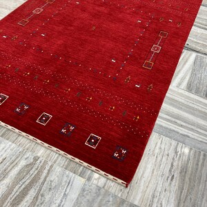 Könnte beinhalten: Ein leuchtend roter Teppich mit geometrischem Muster. Der Teppich hat ein durchgehend rotes Mittelfeld, das mit Reihen kleiner, farbiger Formen und rechteckigen Bordüren verziert ist. Die Ränder sind mit einem cremefarbenen Rand versehen.