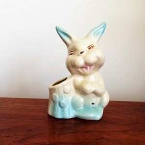 May include: A white ceramic bunny planter with blue ears and a blue and white tree stump base. The bunny has a smiling face and a pink tongue.