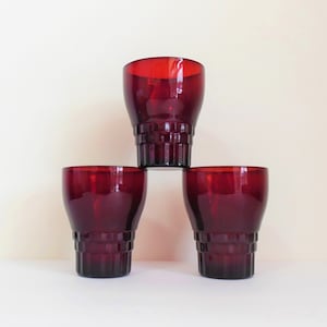 May include: Four ruby red glass tumblers stacked in a pyramid shape. The glasses have a textured, ribbed design.