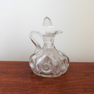 May include: A clear glass bottle with a textured surface and a small spout. The bottle has a decorative design of circles and swirls.