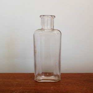 May include: A clear glass bottle with a square body and a short neck. The bottle has a textured surface and is empty.
