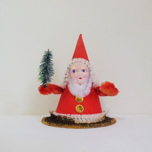 May include: A vintage Christmas decoration featuring a red felt Santa Claus holding a small green tree. The Santa is wearing a white beard and a red hat with white trim. The decoration is sitting on a brown base.