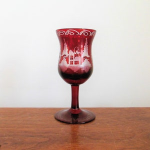 May include: A red glass goblet with a white etched design of a house and trees. The goblet has a round base and a tall, slender stem.