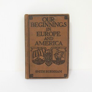 May include: A brown book titled "Our Beginnings in Europe and America" by Smith Burnham. The cover features a decorative design with three shields, each containing a ship and a symbol.