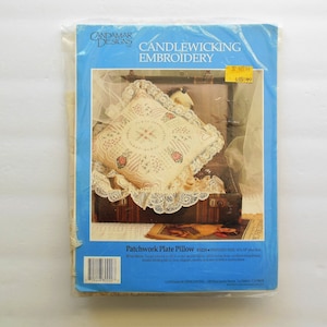 May include: A blue and white pamphlet with the title "Candlewicking Embroidery" and a picture of a white pillow with lace trim. The pillow is on a wooden chest with a book on top. The text "Patchwork Plate Pillow" is visible on the pamphlet.