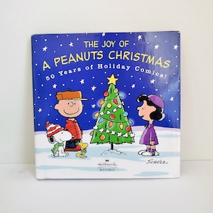 May include: A blue book cover with the title "The Joy of A Peanuts Christmas" and the text "50 Years of Holiday Comics!"  The cover features illustrations of Charlie Brown, Snoopy, Lucy, and a decorated Christmas tree.
