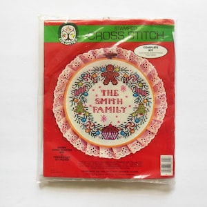 May include: A Colortex stamped cross stitch kit featuring a Christmas wreath with a gingerbread man, candy canes, ornaments, and a cupcake. The wreath is surrounded by a pink lace trim and the text "The Smith Family" is in the center. The kit is complete and includes everything needed to complete the project.