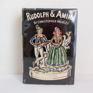 May include: A black book cover with a colorful illustration of a man and woman in traditional clothing. The title of the book is "Rudolph & Amina" by Christopher Morley.