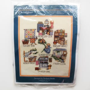 May include: A cross-stitch kit by Bobbie Designs, featuring a colorful sampler with various domestic scenes labeled with days of the week. The finished size is 10 x 13 inches. The text "Women's Work is Never Done" is visible.