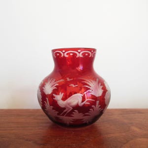 May include: A red glass vase with a white etched design of a deer, trees, and birds. The vase has a rounded shape and a decorative rim. The etched design is detailed and creates a contrast against the red glass.