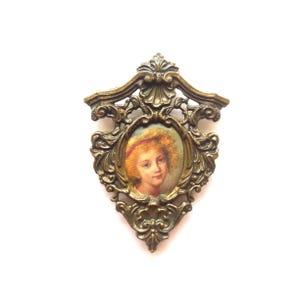 May include: A gold-toned metal brooch with an ornate frame and a cameo of a young girl with blonde hair. The cameo is set in a circular frame with a raised edge.