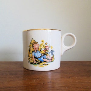 May include: White ceramic mug with a gold rim. The mug features a cartoon illustration of a boy sitting in a pile of hay with a cow beside him.
