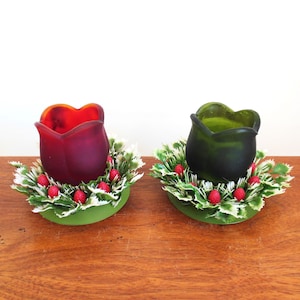 May include: Two glass candle holders in the shape of a tulip, one red and one green, are surrounded by a green and white holly wreath with red berries.