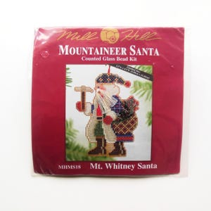 May include: A red package with the text "Mountaineer Santa" and "Counted Glass Bead Kit". The image shows a cross-stitch ornament of Santa Claus holding a hammer and a basket. The finished size is 3.5" x 4.5".