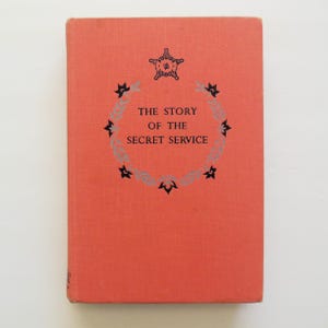 May include: A red book with the title "The Story of the Secret Service" in black lettering. The book cover features a black star with a silver outline and a decorative border of black and silver leaves.