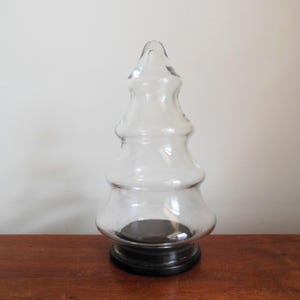 May include: A clear glass Christmas tree shaped jar with a black base. The jar is empty and has a rounded, bulbous shape.